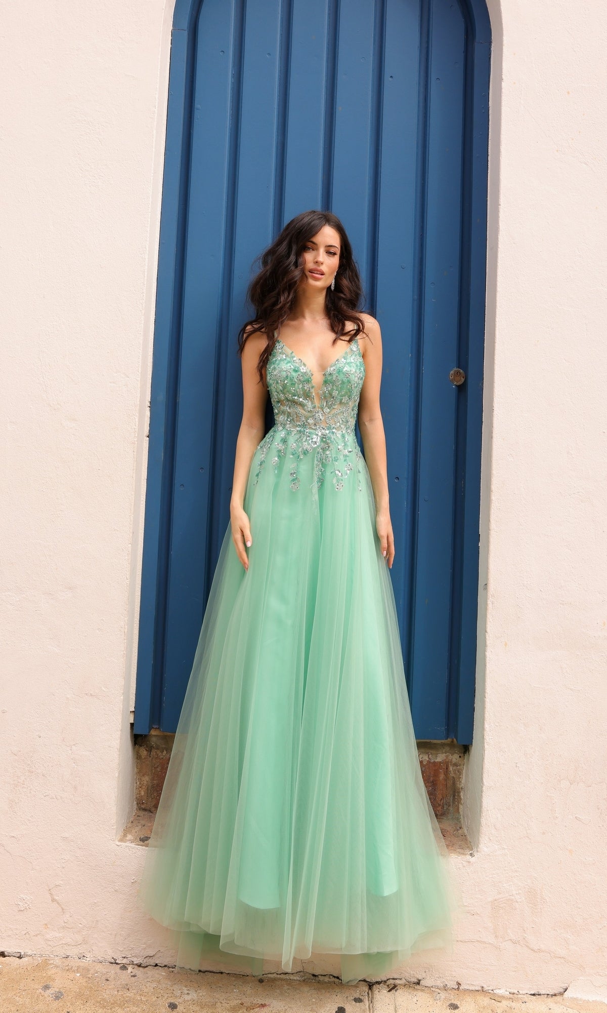 A woman in the Deep V-Back Long Tulle A-Line Prom Dress Q1391, a sleeveless mint green gown adorned with embroidered flowers, stands before a tall blue door framed by a white wall.